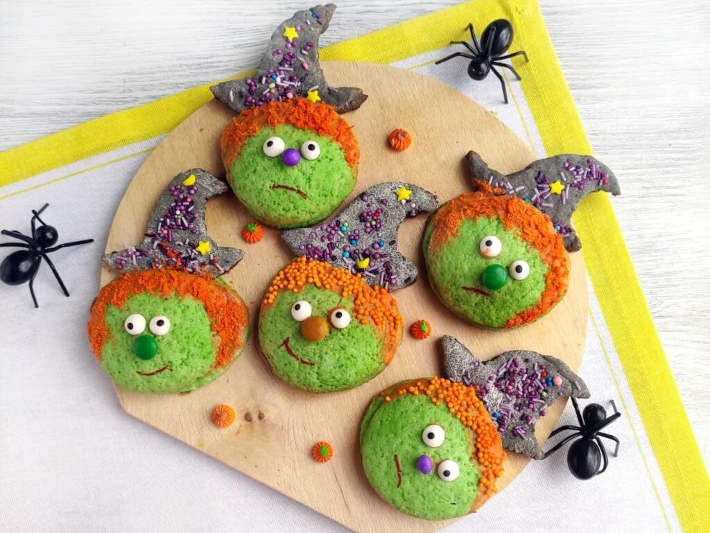 Decorated candy-filled Halloween witch cookies with green faces, orange sprinkle hair, sugar eyes, and witch hats, displayed on a wooden board with plastic spiders for Halloween.