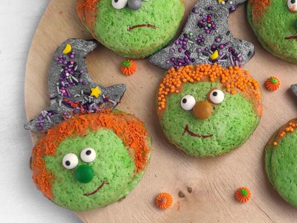 Finished candy-filled Halloween witch cookies decorated with sugar eyes, orange sprinkles for hair, and black witch hats with colorful sprinkles.