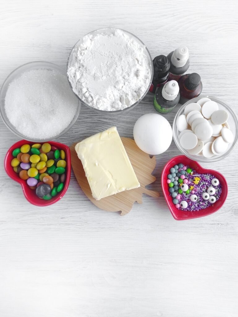 Ingredients for candy-filled Halloween witch cookies including butter, flour, sugar, egg, food coloring, white chocolate, sprinkles, and candy eyes.