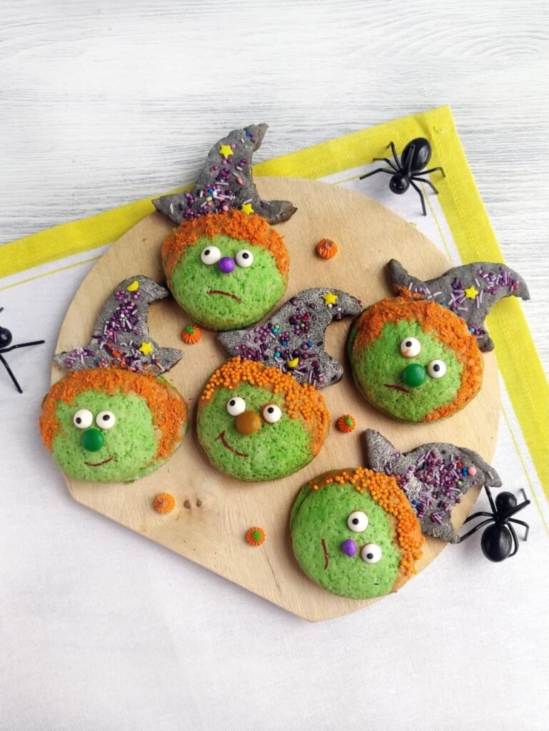 Platter of candy-filled Halloween witch cookies decorated with green faces, orange sprinkle hair, sugar eyes, and witch hats with colorful sprinkles, surrounded by plastic spiders.
