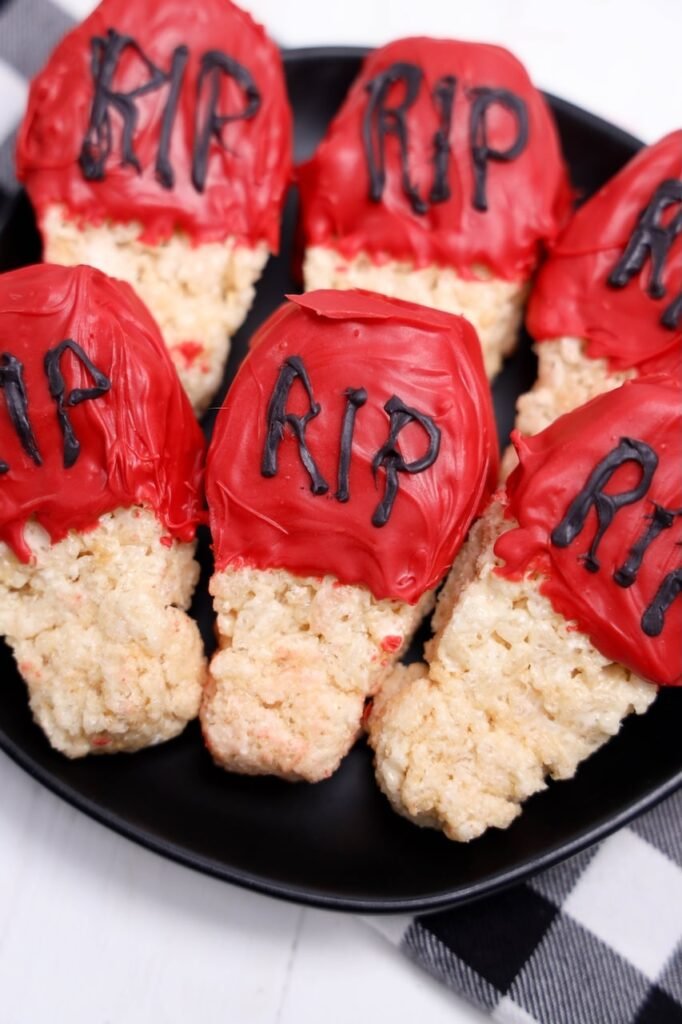 Close-up of coffin-shaped Rice Krispies Halloween treats dipped in red candy melts with black RIP lettering on a black plate.