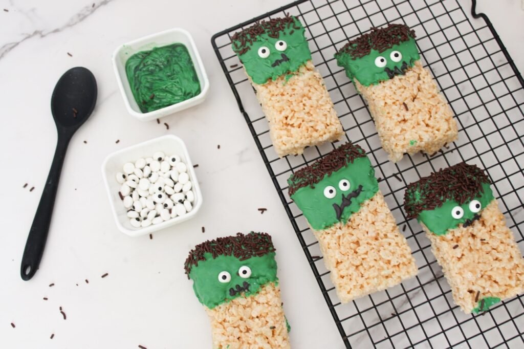 Partially decorated Frankenstein Rice Krispies Treats on a cooling rack with bowls of green candy melts and candy eyes on a white countertop.