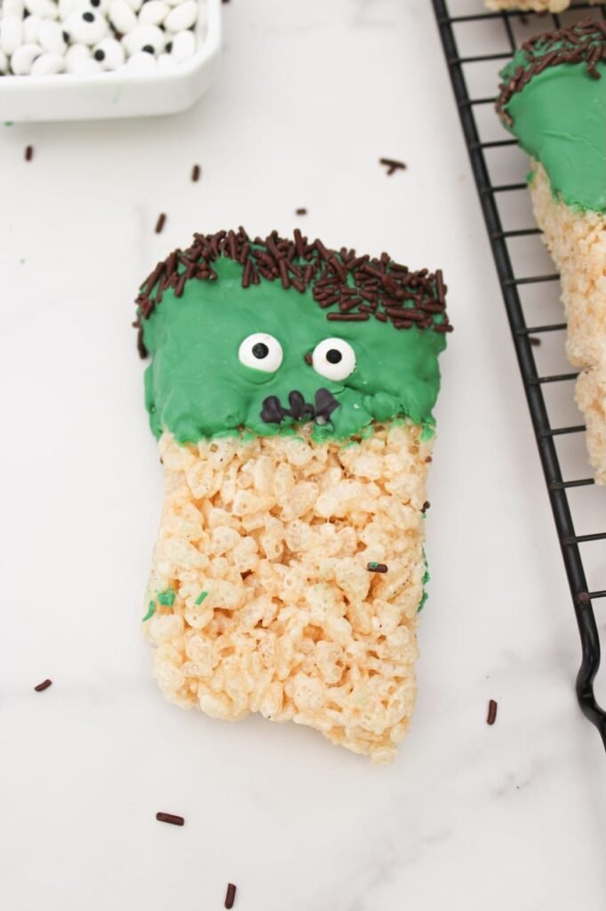 Close-up of a Frankenstein Rice Krispie treat dipped in green chocolate with candy eyes, chocolate sprinkle hair, and a piped black mouth, resting on a white marble surface.