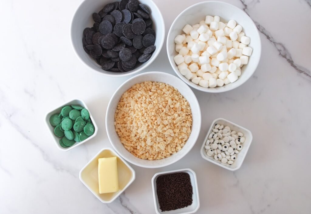 Ingredients for Frankenstein Rice Krispie Treats including Rice Krispies cereal, marshmallows, butter, green candy melts, black chocolate melts, candy eyes, and chocolate sprinkles arranged in bowls