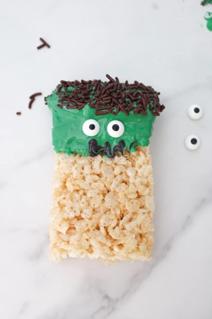 Finished Frankenstein Rice Krispies Treat with green candy coating, chocolate sprinkle hair, candy eyes, and black icing mouth, displayed on a white marble surface.