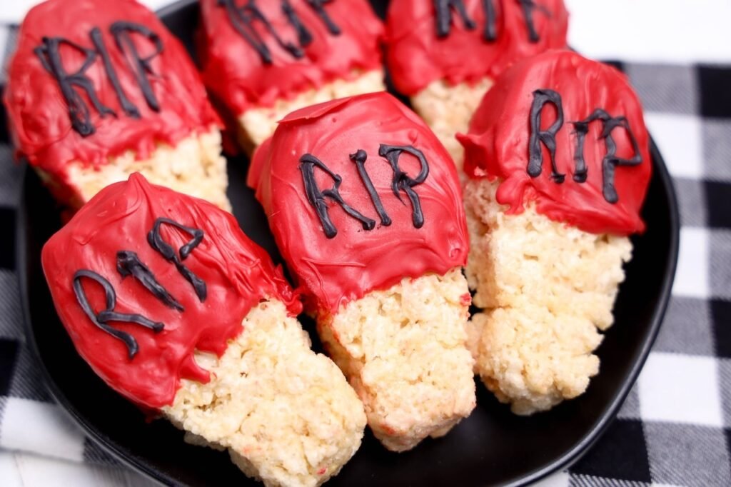 Coffin-shaped Halloween Rice Krispies treats dipped in red candy melts with RIP piped in black chocolate, served on a black plate.