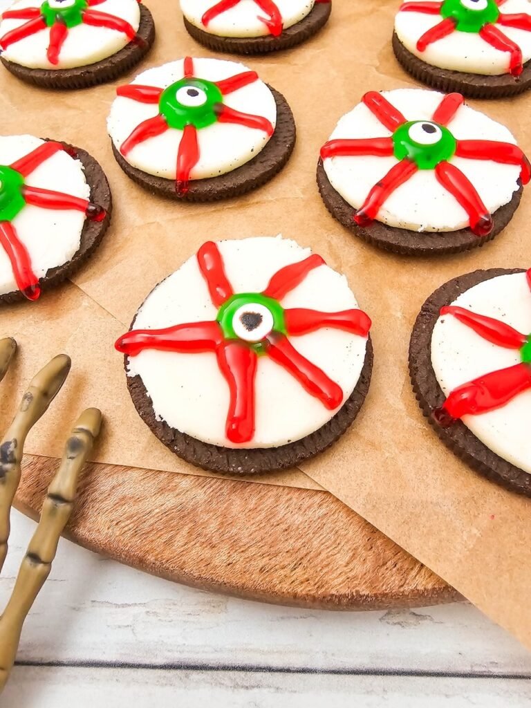 Several decorated Oreo Eyeballs arranged on parchment paper over a wooden serving board, featuring Oreo cookies topped with green candy eyeballs and red icing veins for a spooky Halloween party treat.