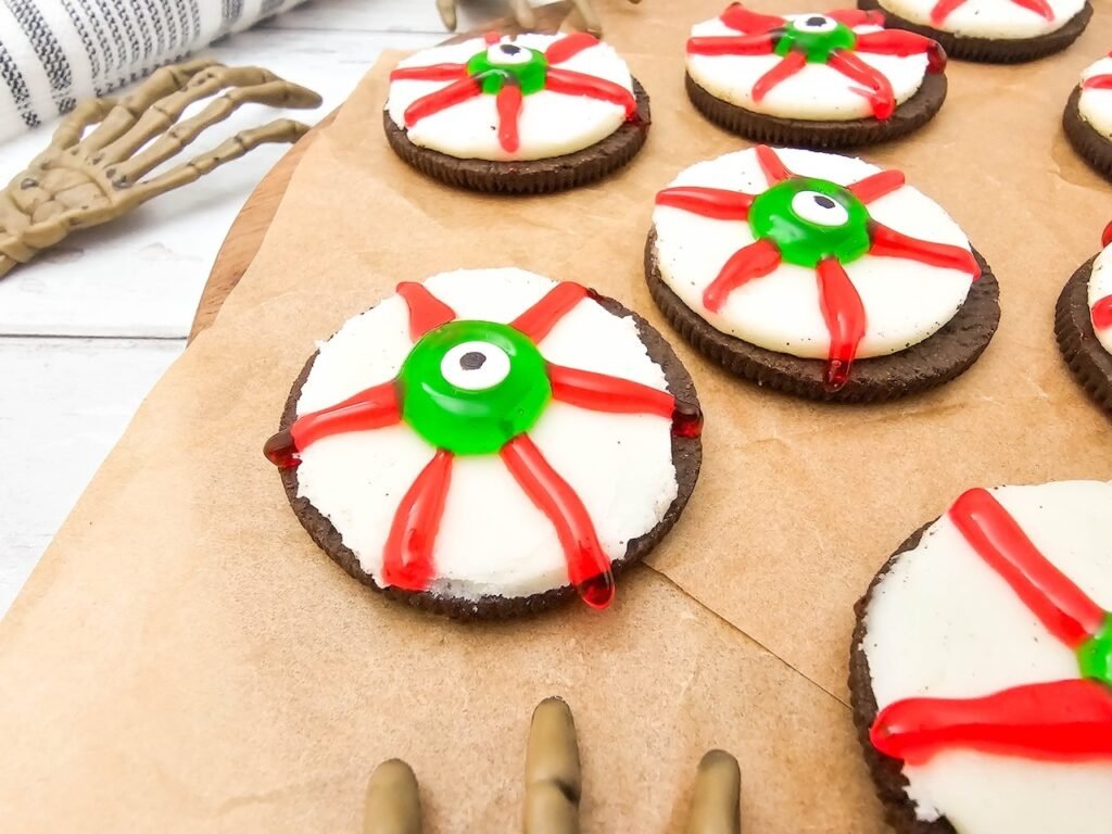 Close-up of decorated Oreo Eyeballs arranged on parchment paper, featuring Double Stuf Oreo cookies topped with green candy eyeballs and red icing veins to create a spooky Halloween dessert.