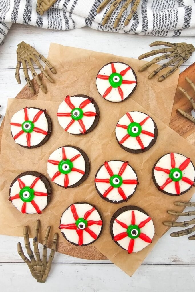 Overhead view of Oreo Eyeballs arranged on parchment paper atop a wooden board, decorated with green candy eyeballs and red icing veins, surrounded by Halloween skeleton hand decorations.
