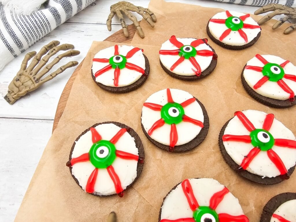 Halloween Oreo eyeball cookies decorated with green candy eyes and red gel veins on a wooden board with skeleton hand props.