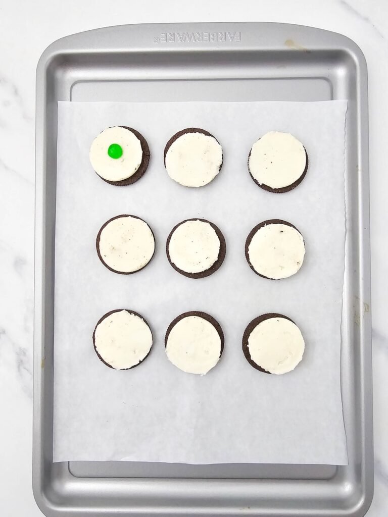 cookies with cream filling on a baking sheet, one cookie topped with a green candy pupil.