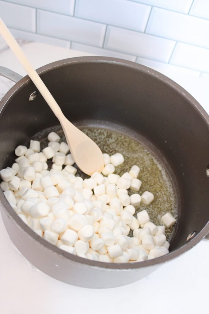 A large saucepan on a white countertop with melted butter and mini marshmallows being stirred with a wooden spoon, showing the beginning steps of making homemade Rice Krispie treats before adding the cereal.