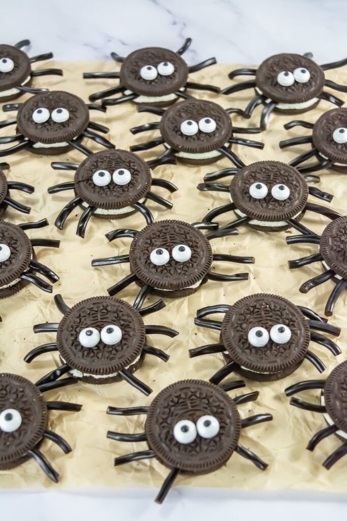 Close-up of Oreo Spider Cookies with black licorice legs and candy eyeballs arranged on parchment paper, a fun no-bake Halloween cookie recipe for kids.