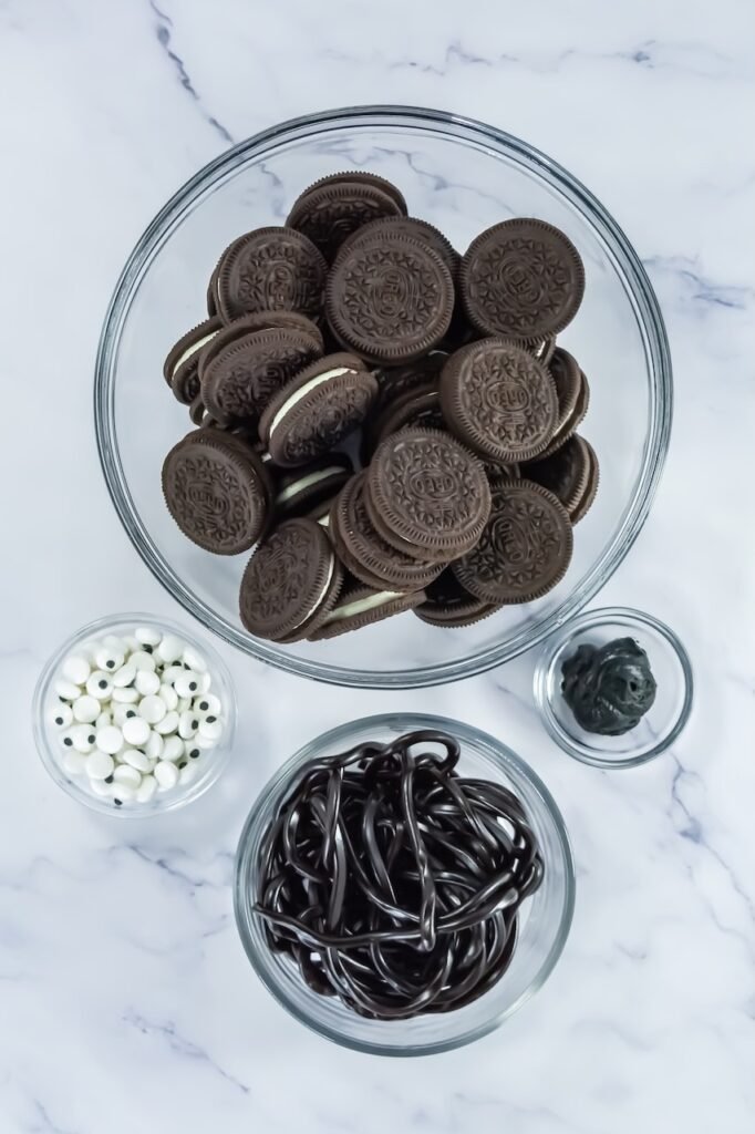 Ingredients for Oreo Spider Cookies on a marble surface including Oreos, licorice ropes, candy eyeballs, and black icing in glass bowls.
