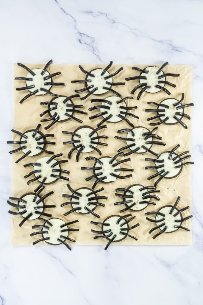 Oreo cookies with cream filling arranged on parchment paper, each with eight black licorice pieces pressed into the filling to create spider legs.