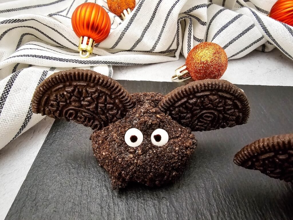 Close-up of a finished Oreo Truffle Bat with Oreo cookie halves for wings and candy eyeballs on a black slate board, with striped cloth and orange decorations in the background, a fun no bake Halloween dessert.