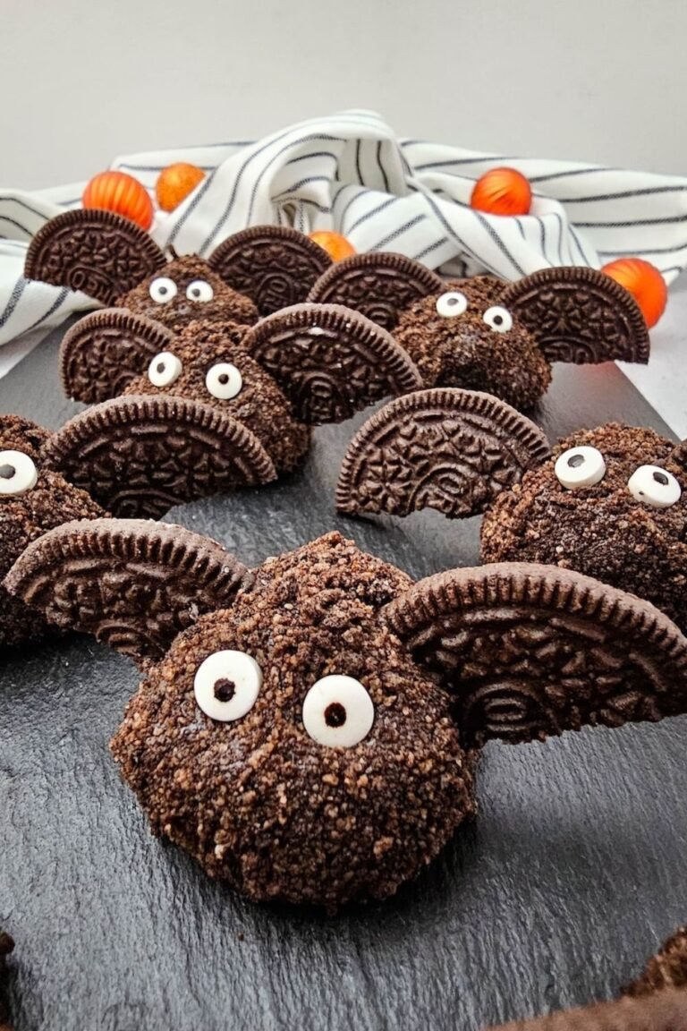 Close-up of a finished Oreo Truffle Bat with Oreo cookie halves for wings and candy eyeballs on a black slate board, with striped cloth and orange decorations in the background, a fun no bake Halloween dessert.