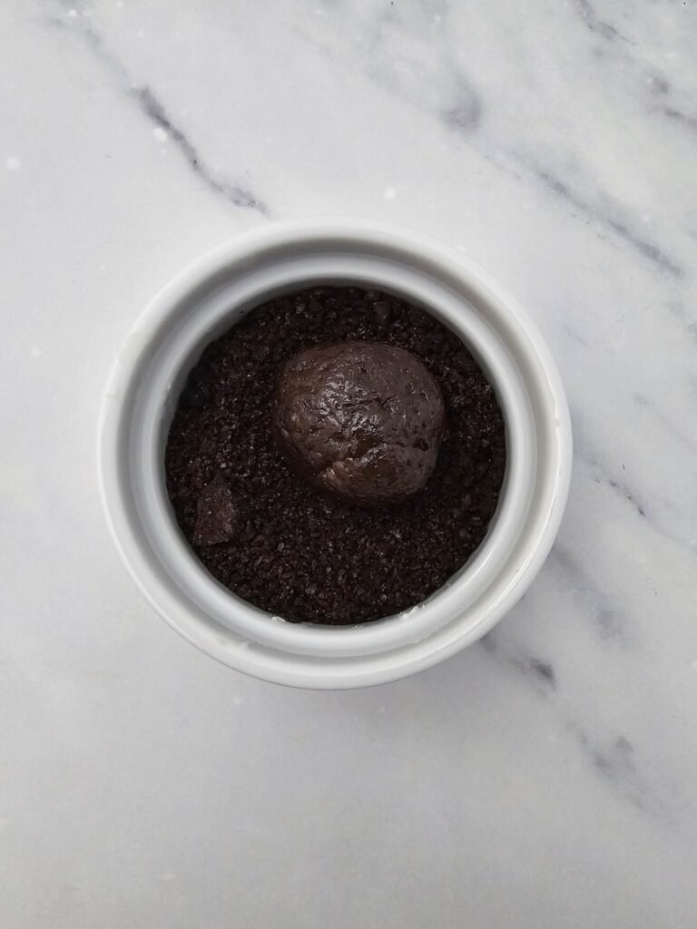 Chocolate Oreo truffle ball placed in a small white bowl filled with crushed Oreo crumbs, being rolled and coated to prepare for decorating Oreo Truffle Bats no bake Halloween treats.