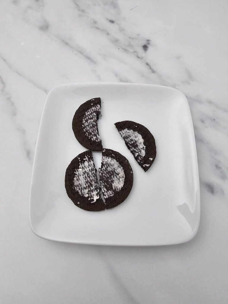 Close-up of two Oreo cookies split in half and broken into smaller pieces on a square white plate, showing how to prepare the cookie halves that will be used as bat wings.