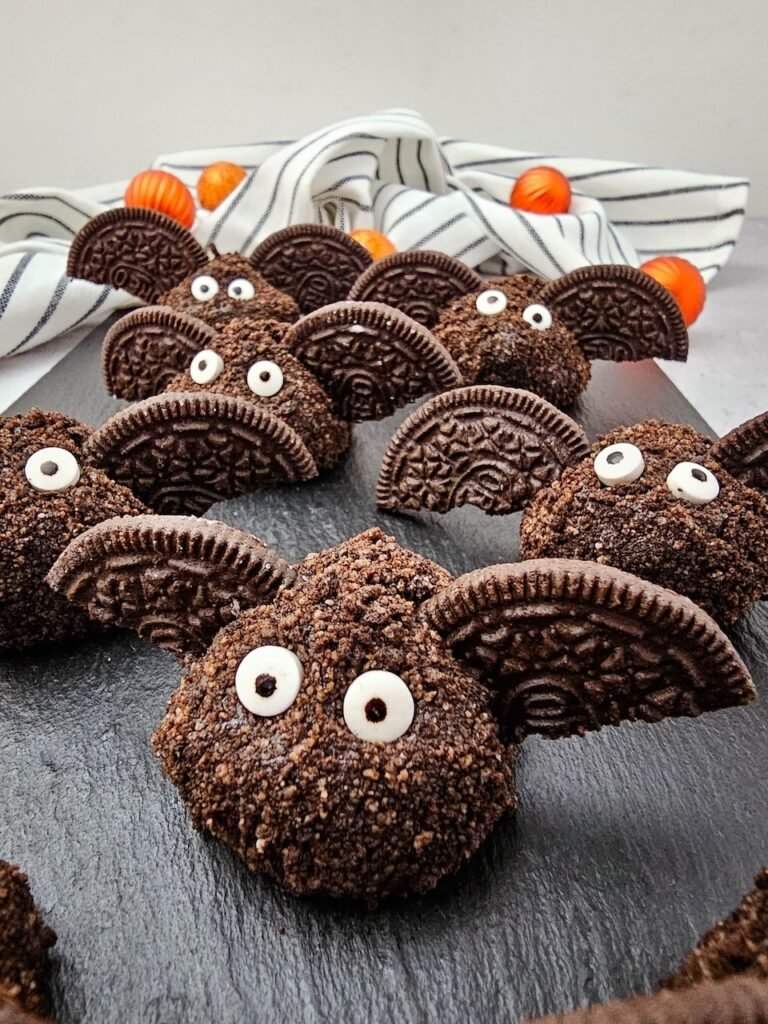 Close-up of Oreo Truffle Bats arranged on a black slate serving board, made with Oreo cookie truffles coated in crumbs, Oreo cookie halves for wings, and candy eyes for a spooky no bake Halloween treat.