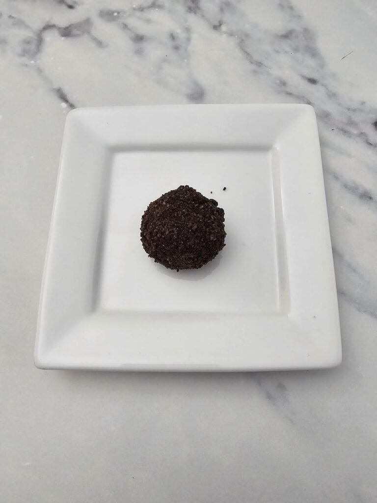 single Oreo truffle ball coated in fine crushed Oreo crumbs and placed on a square white plate, ready to be decorated with wings and candy eyes to create Oreo Truffle Bats Halloween treats.