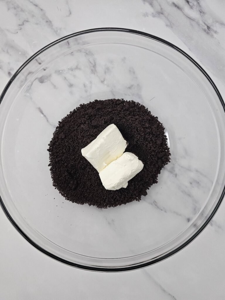 Glass mixing bowl filled with fine Oreo cookie crumbs and two blocks of cream cheese on top, ready to be blended together to form the filling for no bake Halloween treats.