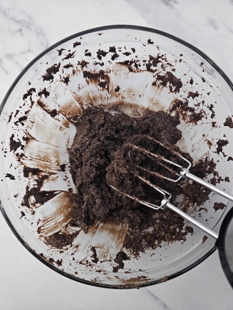 Glass bowl with Oreo crumbs and cream cheese fully mixed into a thick chocolate dough using a hand mixer, creating the base filling for Oreo Truffle Bats no bake Halloween treats.