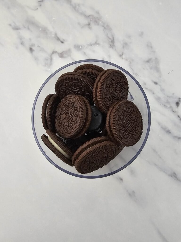 Whole Oreo cookies placed inside a food processor bowl on a marble counter, ready to be crushed into fine crumbs for making.