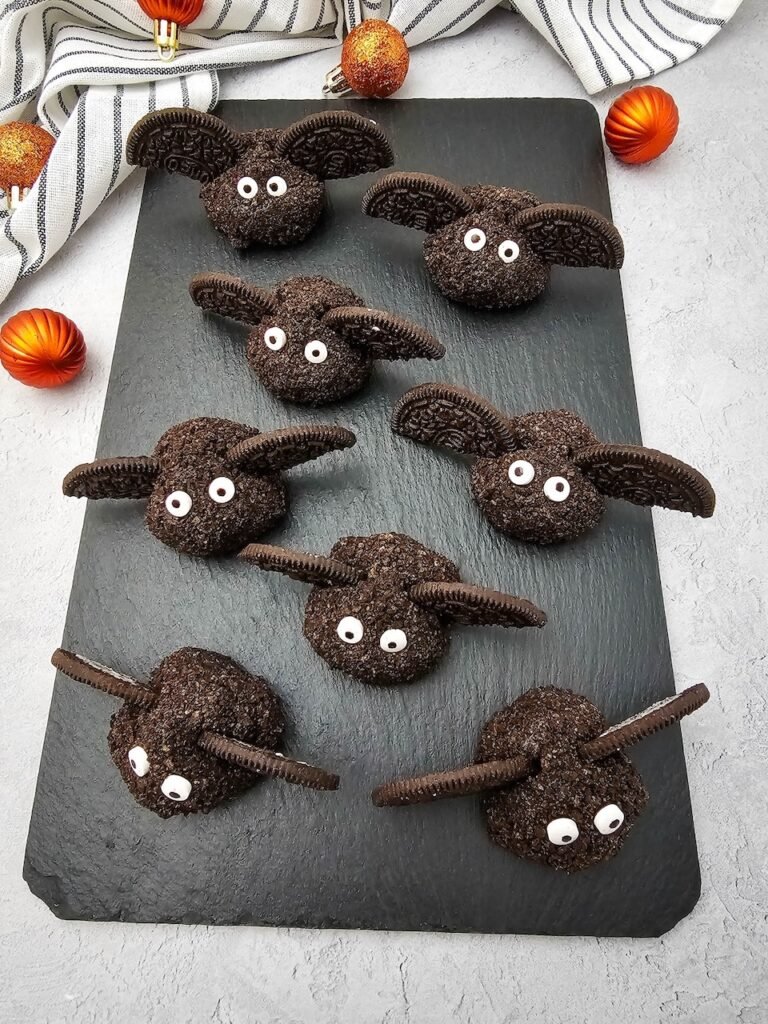 Eight Oreo Truffle Bats arranged neatly on a black slate tray with Oreo cookie wings and candy eyes, served as a festive no bake Halloween party dessert with striped cloth and orange decorations in the background.