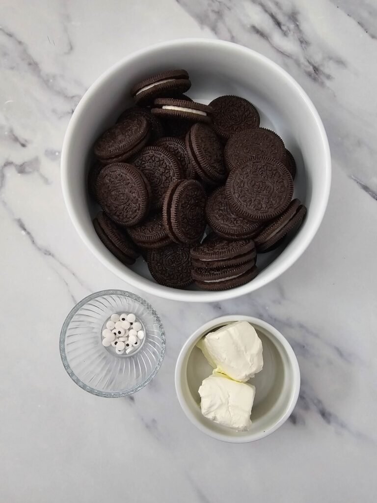 Baking ingredients laid out on a marble countertop including a white bowl filled with Oreo cookies, a small glass dish with candy eyeballs, and a small ceramic bowl with two blocks of cream cheese for making no bake dessert.