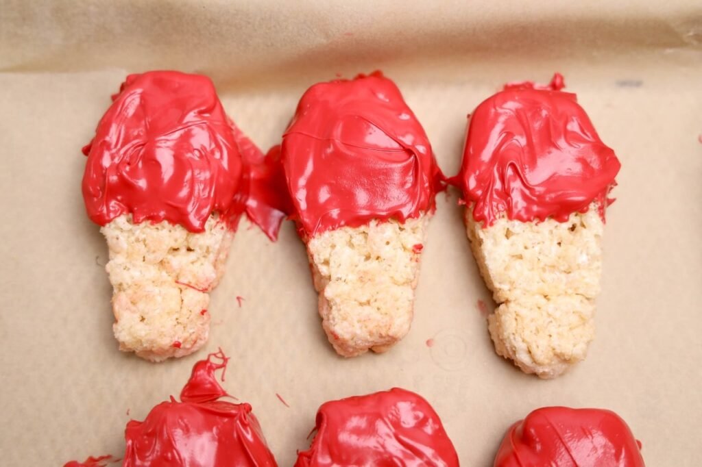 Coffin-shaped Rice Krispies treats dipped halfway in red candy melts setting on parchment paper for Halloween desserts.