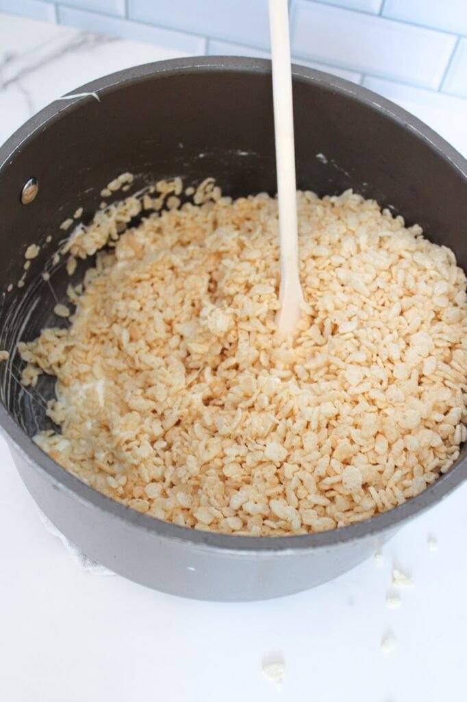 A large saucepan filled with Rice Krispies cereal being stirred into melted marshmallows with a wooden spoon, creating the sticky base mixture for Rice Krispie treats.
