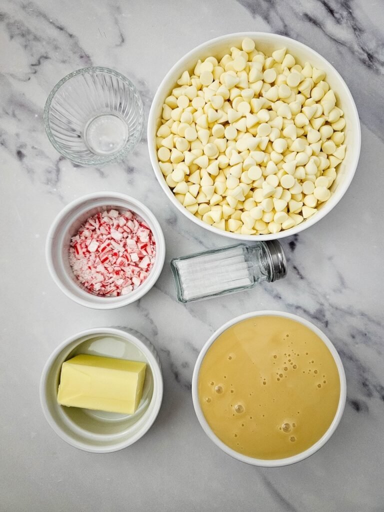 Overhead view of white chocolate chips, sweetened condensed milk, butter, peppermint, and crushed candy canes in small bowls.