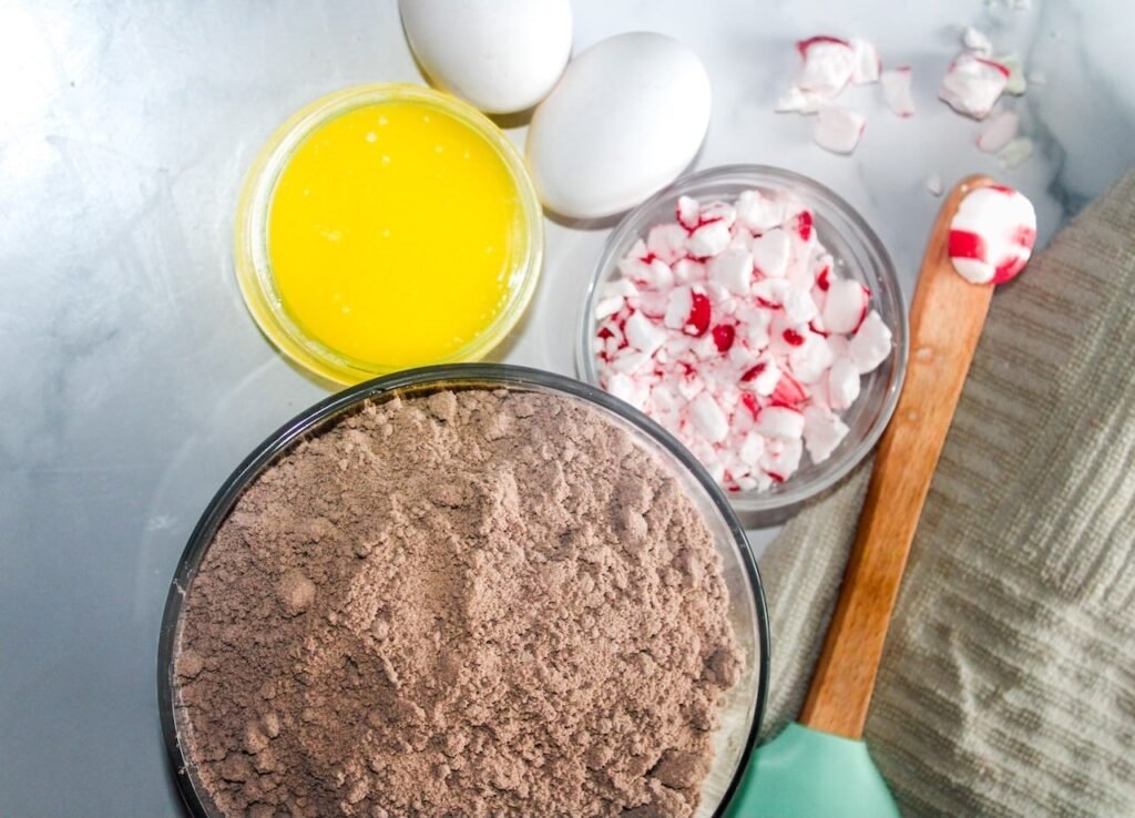 A flat lay of peppermint bark cookie ingredients including chocolate cake mix, crushed peppermint candies, melted butter, and eggs on a kitchen counter.