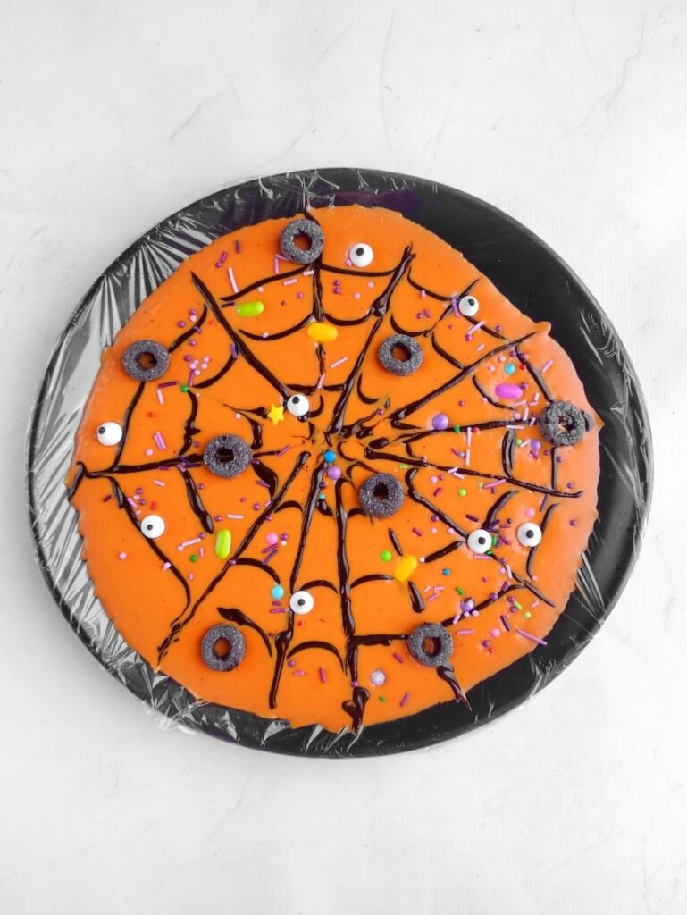 Finished Halloween bark step showing spooky spiderweb design with sugar eyeballs, colorful sprinkles, and candy decorations on orange chocolate.