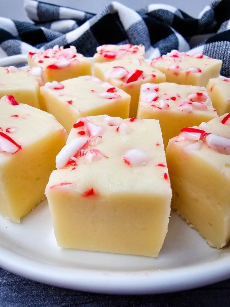 Close-up photo of homemade candy cane fudge made with white chocolate and topped with crushed peppermint candy canes, arranged in even squares on a white plate with a black and white checkered cloth in the background — a festive no-bake Christmas dessert.