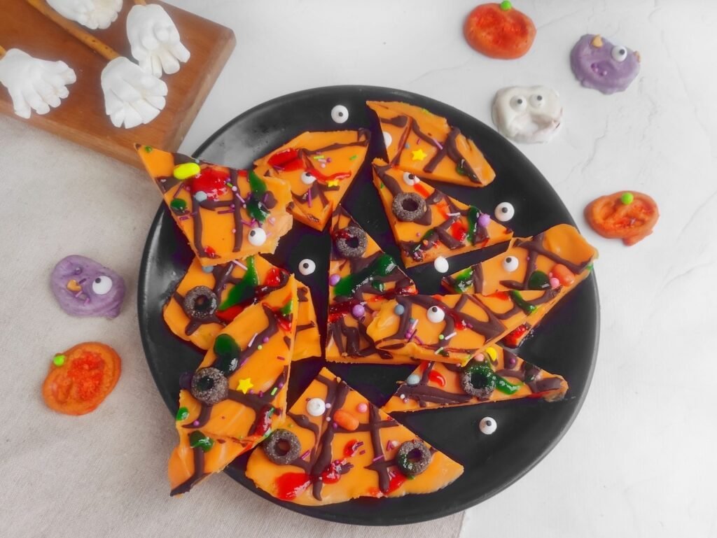 Homemade Halloween candy bark displayed on a black plate, decorated with edible eyes, sprinkles, and colorful toppings surrounded by festive Halloween candies.