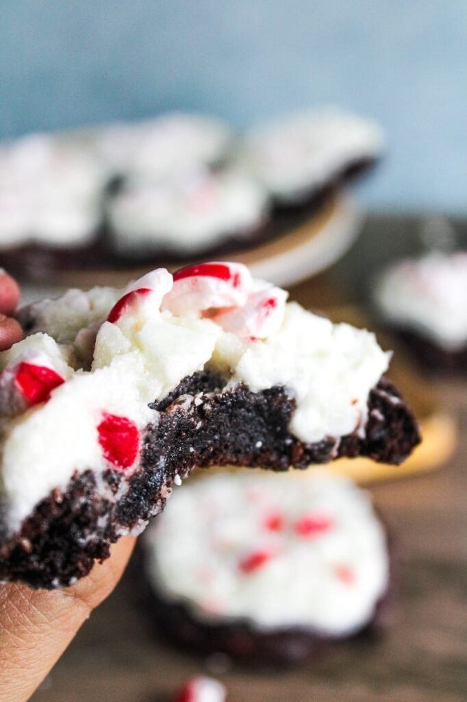 A close-up of a bitten peppermint bark cookie showing a fudgy chocolate center topped with creamy white chocolate frosting and crushed peppermint.