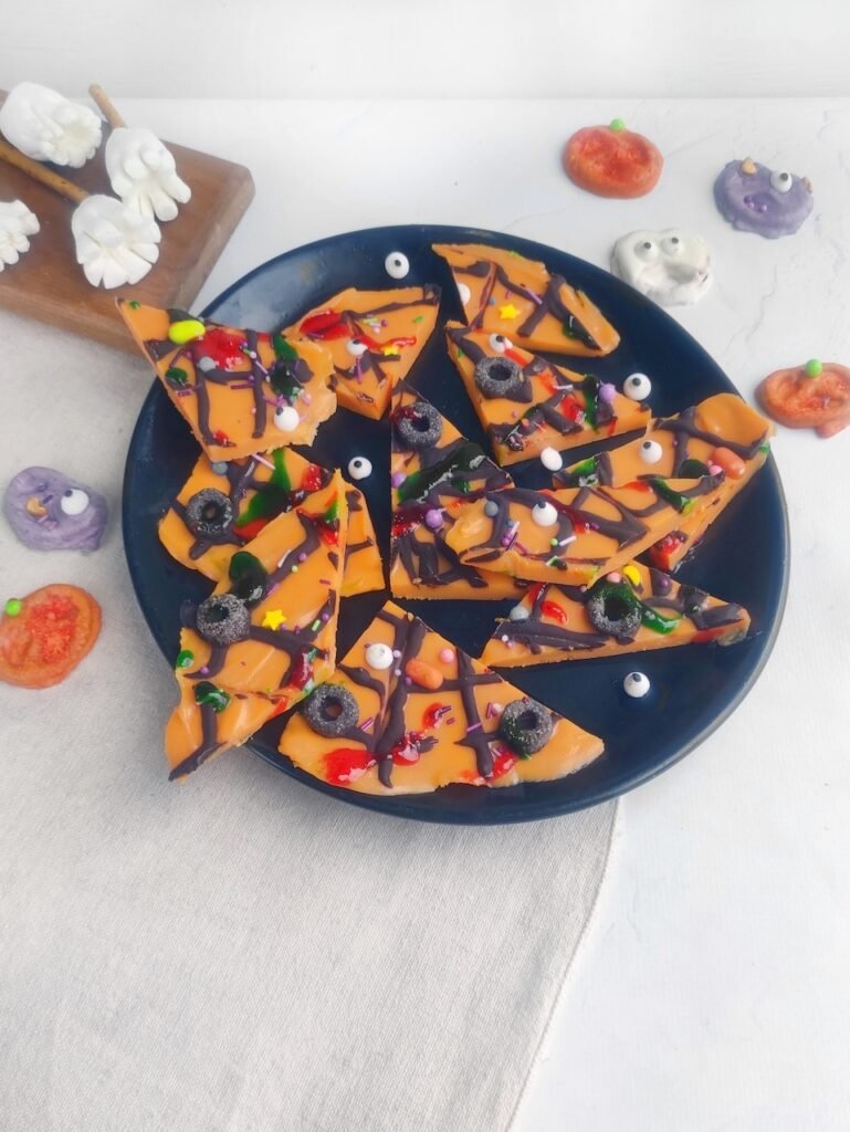 Final step of recipe showing broken pieces of orange chocolate decorated with dark chocolate spiderweb, sugar eyes, sprinkles, and jam drips on a plate.