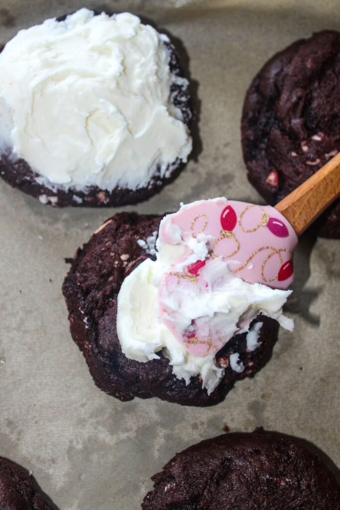 A spatula spreading creamy white chocolate peppermint frosting onto freshly baked chocolate peppermint cookies on a parchment-lined pan.
