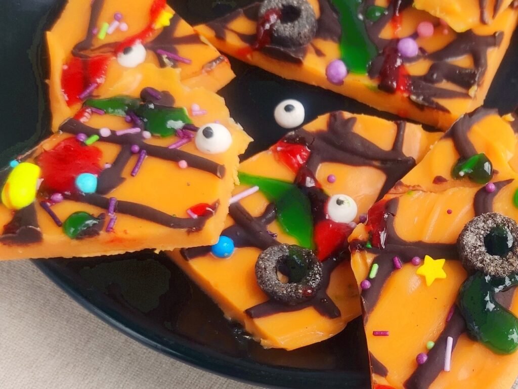 Close-up of dessert showing orange chocolate shards decorated with dark chocolate web drizzle, sugar eyeballs, sprinkles, and colorful candy pieces.