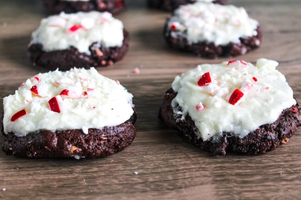 Soft chocolate peppermint bark cookies topped with creamy white frosting and crushed peppermint candies, arranged on a wooden table.