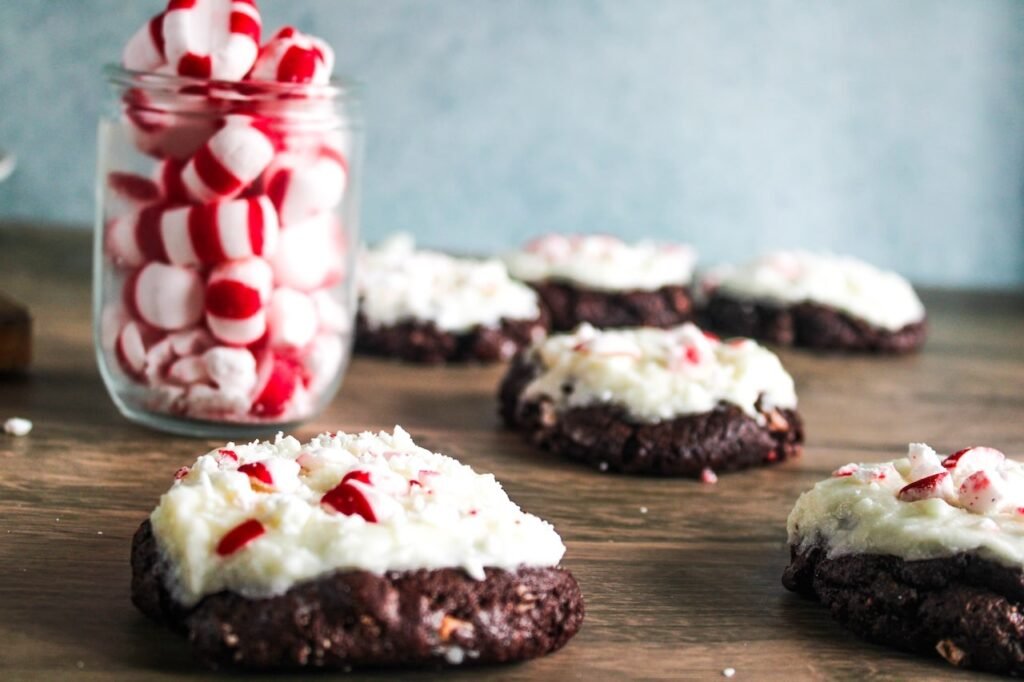 Chocolate peppermint bark cookies topped with creamy white chocolate frosting and crushed peppermint, arranged on a wooden surface beside a jar of peppermint candies.