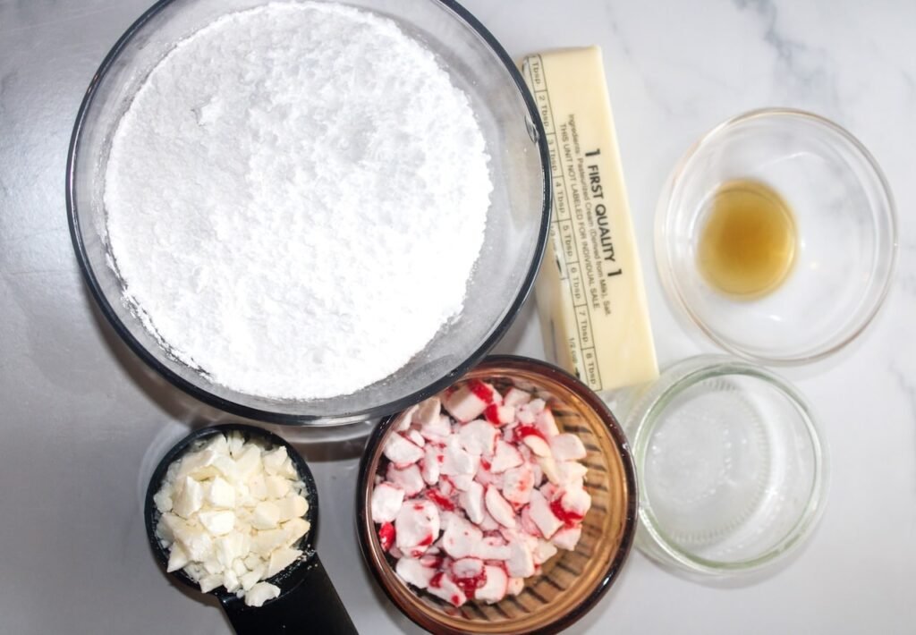 Ingredients for white chocolate peppermint frosting including butter, powdered sugar, peppermint candies, chopped white chocolate, and vanilla extract arranged on a counter.