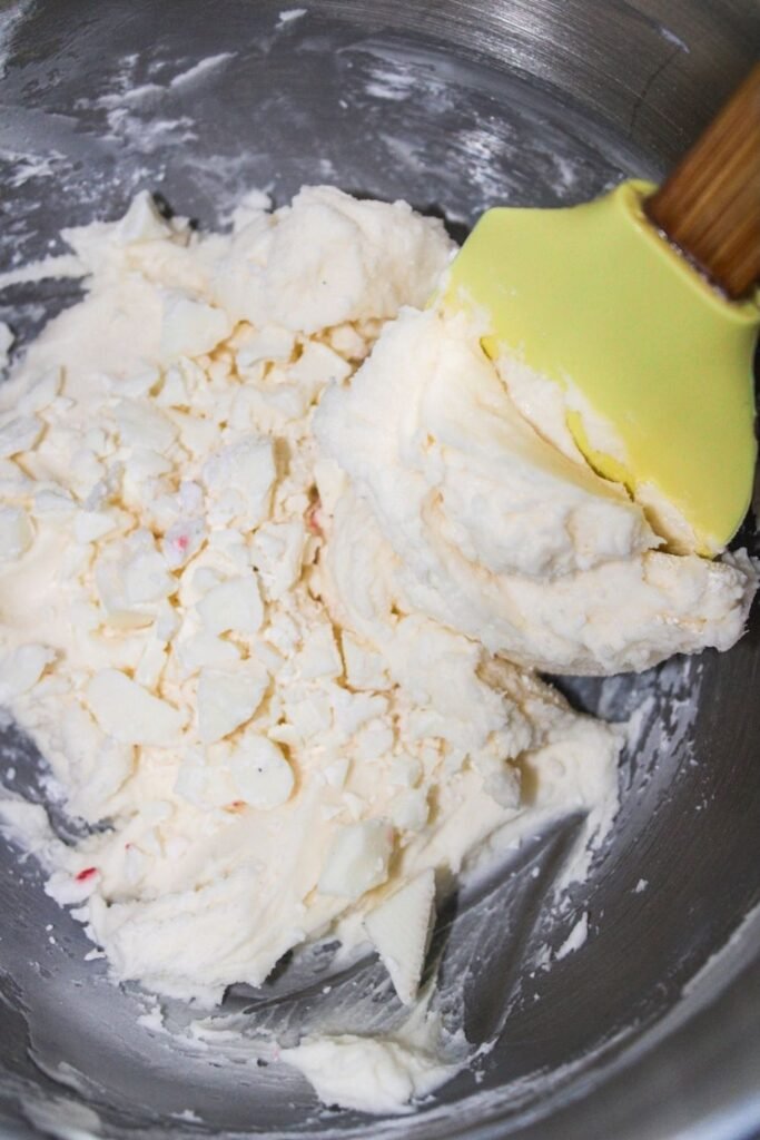 A close-up of creamy white chocolate peppermint frosting being mixed with a spatula in a stainless steel bowl.