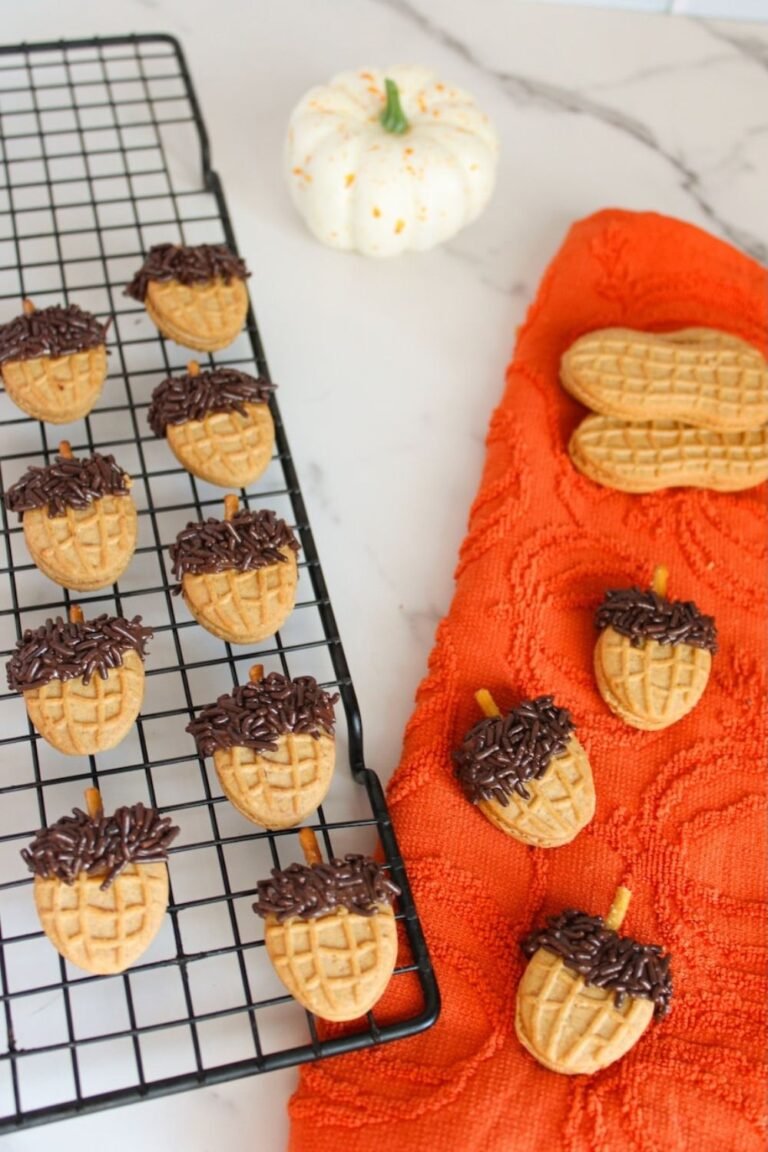 Overhead view of finished Nutter Butter acorn cookies topped with chocolate sprinkles and pretzel stick stems cooling on a wire rack beside an orange fall towel and white pumpkin, styled as an easy no-bake fall dessert.