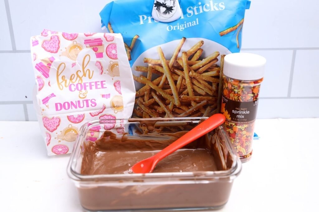 ingredients for acorn donut holes including chocolate melting wafers, pretzel sticks, fall sprinkles, and donut holes on a white counter.