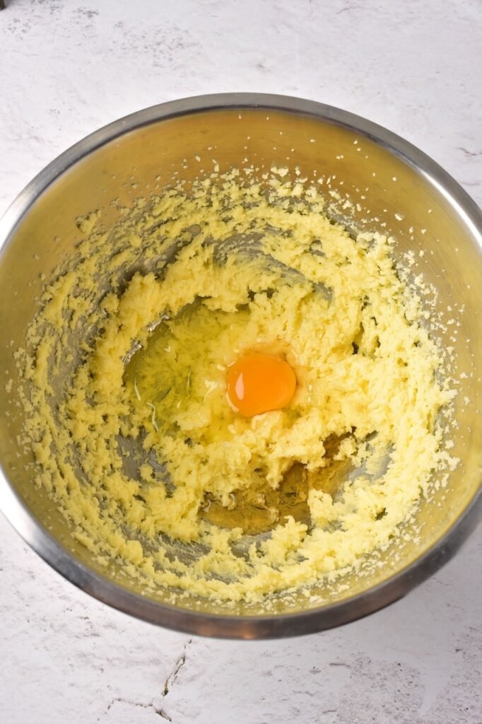 Adding an egg to creamed butter and sugar mixture in a stainless steel bowl.