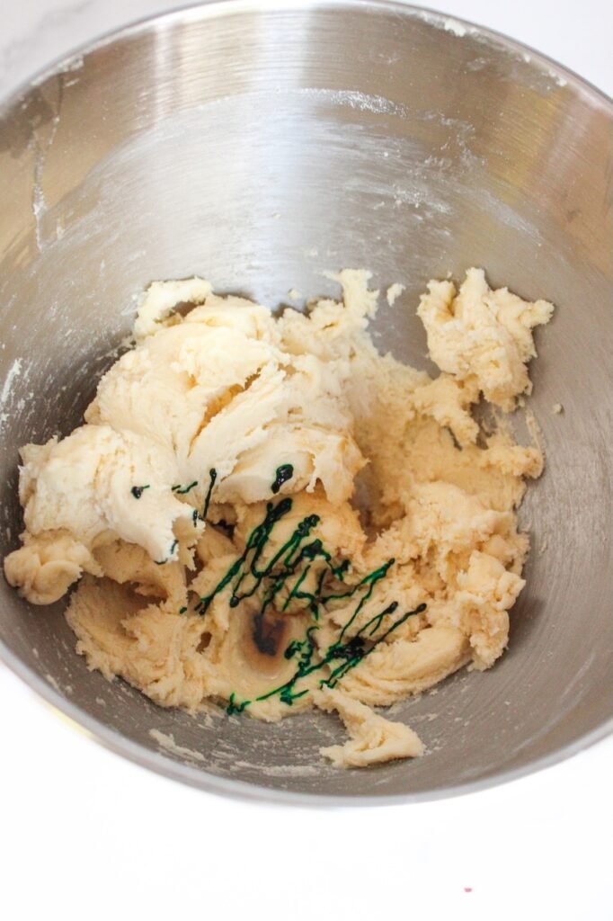 Vanilla cookie dough in a mixing bowl with green food coloring and peppermint extract being added.