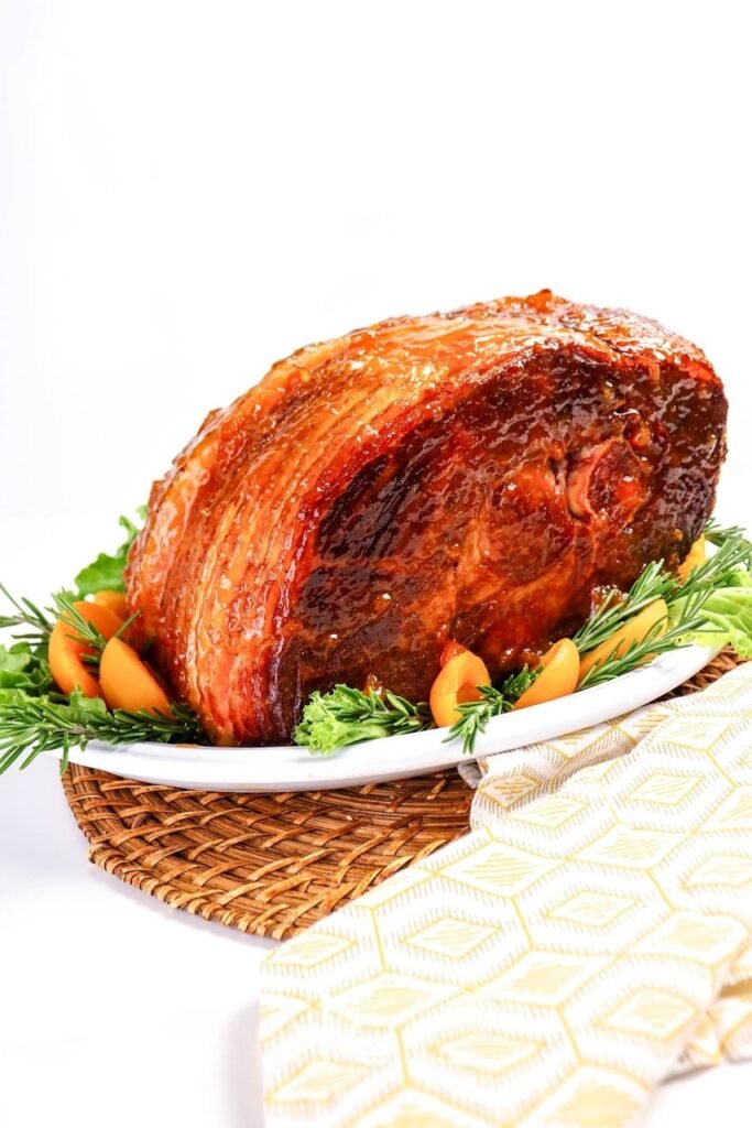 Whole apricot glazed ham with shiny golden crust on a white platter, garnished with apricots, rosemary, and greens — an easy baked holiday ham perfect for Easter or Christmas dinner.